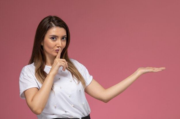 Front view young female in white shirt showing silence sign on the pink wall