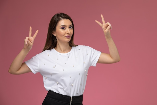 Front view young female in white shirt showing victory sign on pink wall, color model woman pose