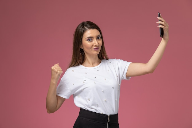 Front view young female in white shirt taking a selfie on the pink wall, model woman