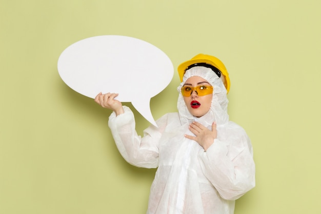 Front view young female in white special suit and yellow helmet holding large white sign on the green space chemistry work