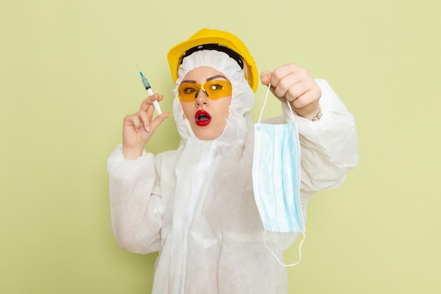 Front view young female in white special suit and yellow helmet holding sterile mask on the green space  