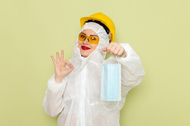 Front view young female in white special suit and yellow helmet holding sterile mask smiling on the green space  