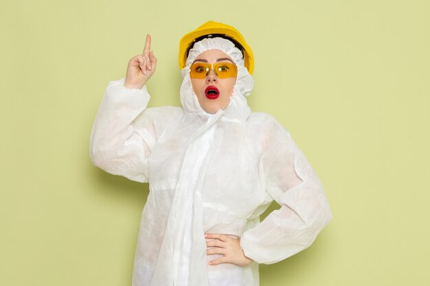 Front view young female in white special suit and yellow helmet posing with raised finger on the green space job 