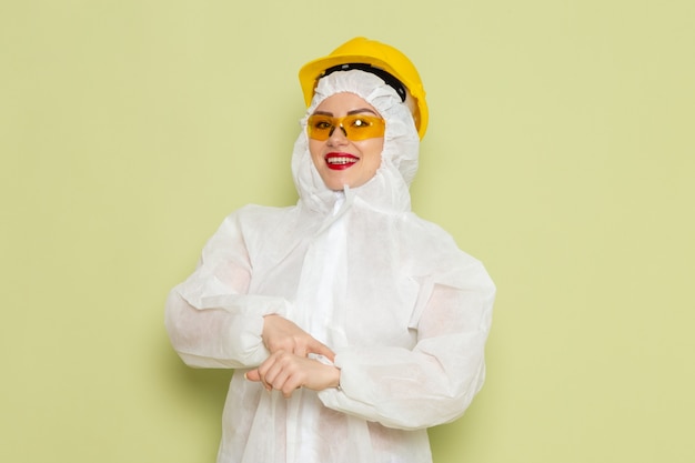 Front view young female in white special suit and yellow helmet smiling pointing out into her wrist on the green space s