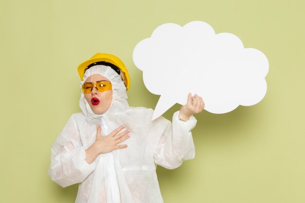 Front view young female in white special suit and yellow protective helmet holding huge white sign on the green space 