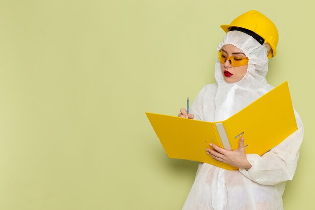 Front view young female in white special suit and yellow protective helmet holding yellow files on the green space suit uniform science