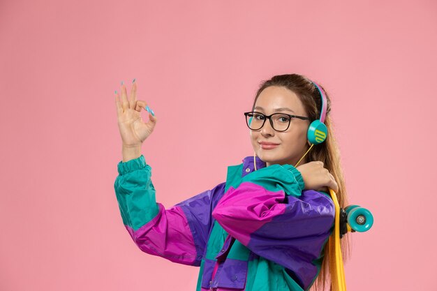 Front view young female in white t-shirt ed coat listening to music via earphones holding skateboard on pink background