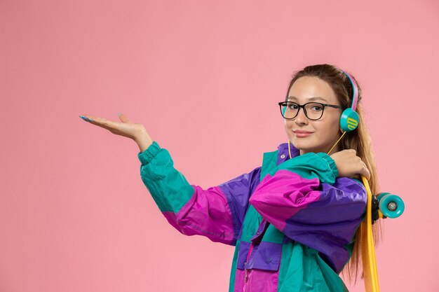 Front view young female in white t-shirt ed coat listening to music via earphones holding skateboard on the pink background