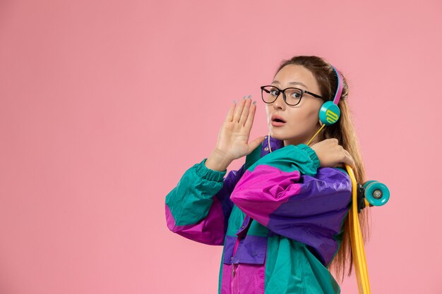 Front view young female in white t-shirt ed coat listening to music via earphones holding skateboard on pink desk female