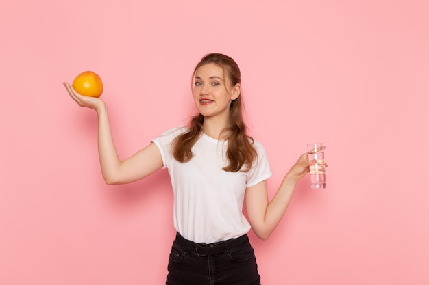 Вид спереди молодой женщины в белой футболке, держащей свежий грейпфрут и стакан воды на розовой стене