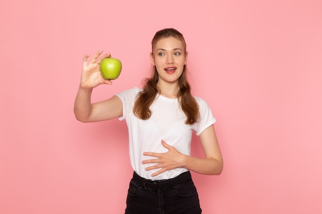 Вид спереди молодой женщины в белой футболке, держащей свежее зеленое яблоко на светло-розовой стене