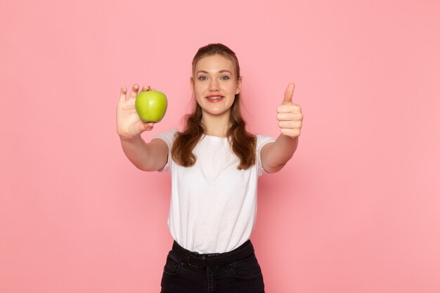 Front view of young female in white t-shirt holding fresh green apple on light pink wall
