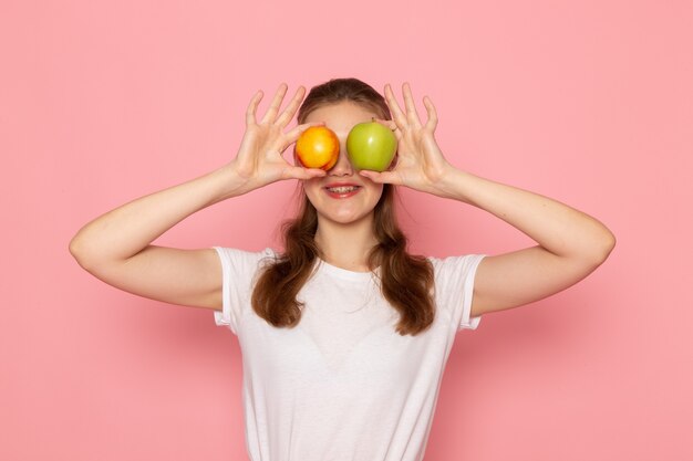 Front view of young female in white t-shirt holding fresh green apple and peach