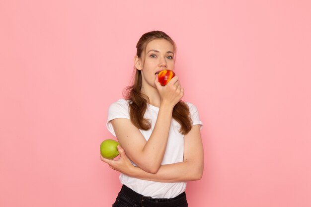 밝은 분홍색 벽에 복숭아 먹는 신선한 녹색 사과를 들고 흰색 티셔츠에 젊은 여성의 전면보기