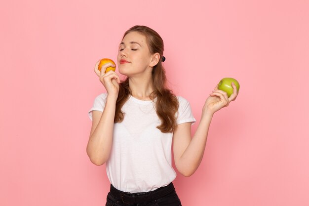 Front view of young female in white t-shirt holding fresh green apple with peach on light-pink wall