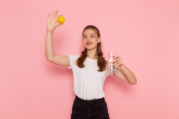 Front view of young female in white t-shirt holding fresh lemon and glass of water on light-pink wall