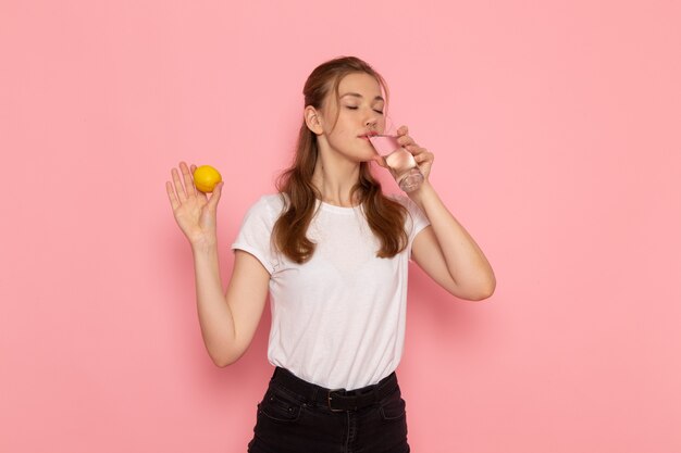 Front view of young female in white t-shirt holding fresh lemon and glass of water on the pink wall