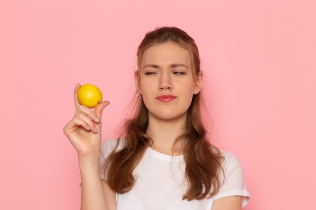 Front view of young female in white t-shirt holding fresh lemon on the lightpink wall