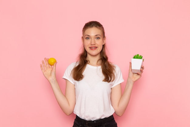 Front view of young female in white t-shirt holding fresh lemon little plant on pink wall