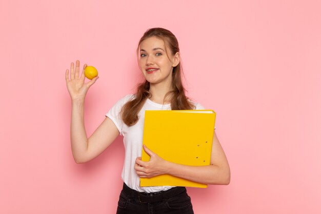 ピンクの壁にファイルと新鮮なレモンを保持している白いTシャツの若い女性の正面図