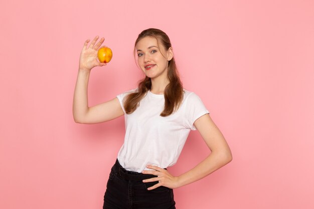 Front view of young female in white t-shirt holding fresh peach on light-pink wall