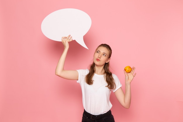 Front view of young female in white t-shirt holding fresh peach and white sign on light-pink wall