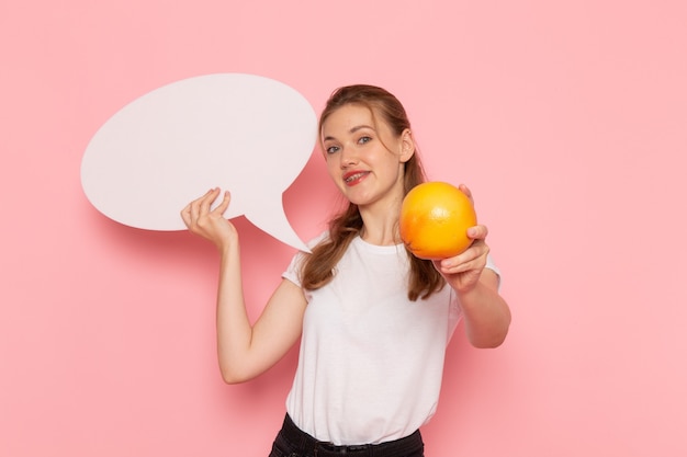 Front view of young female in white t-shirt holding grapefruit and white sign smiling on pink wall