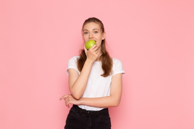 Вид спереди молодой женщины в белой футболке, держащей зеленое яблоко, кусающей его на розовой стене