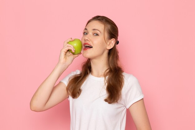 ピンクの壁にそれを食べる青リンゴを保持している白いTシャツの若い女性の正面図