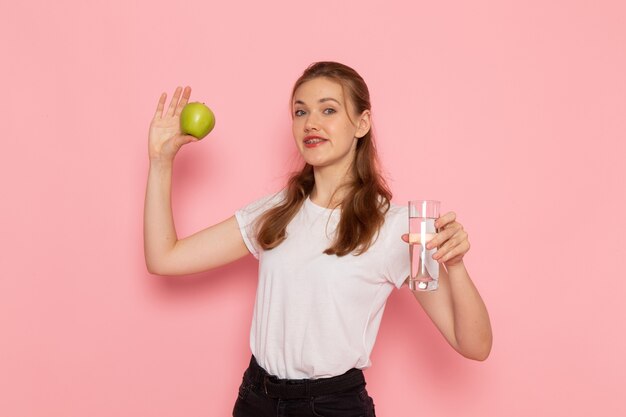 Front view of young female in white t-shirt holding green apple and glass of water on pink wall