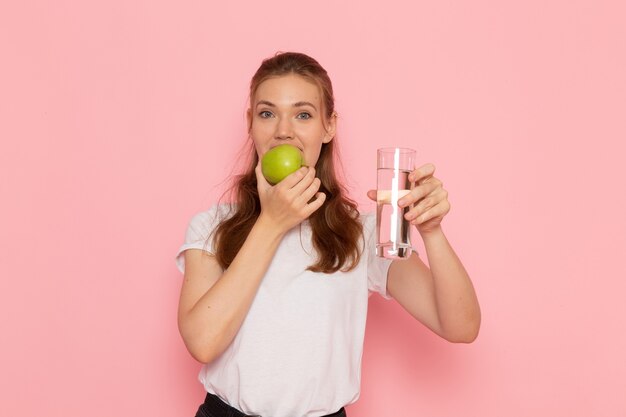 Front view of young female in white t-shirt holding green apple and glass of water on the pink wall