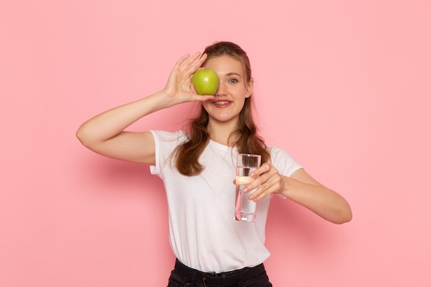 緑のリンゴとピンクの壁に笑みを浮かべて水のガラスを保持している白いTシャツの若い女性の正面図