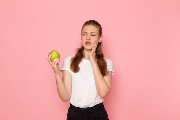 Вид спереди молодой женщины в белой футболке, держащей зеленое яблоко с зубной болью на розовой стене