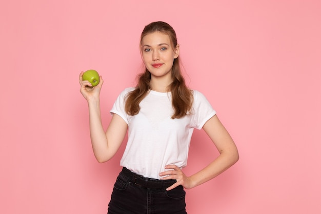 Front view of young female in white t-shirt holding green apple on pink wall