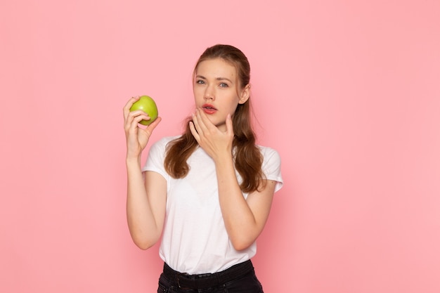 Front view of young female in white t-shirt holding green apple and thinking on pink wall