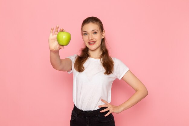 Front view of young female in white t-shirt holding green apple with smile on light pink wall