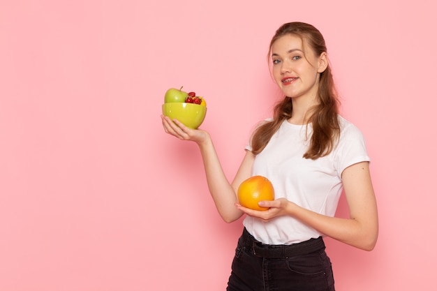 Front view of young female in white t-shirt holding plate with fresh fruits and grapefruit on the pink wall