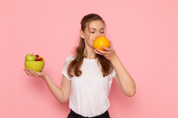 Front view of young female in white t-shirt holding plate with fresh fruits and grapefruit smelling on pink wall