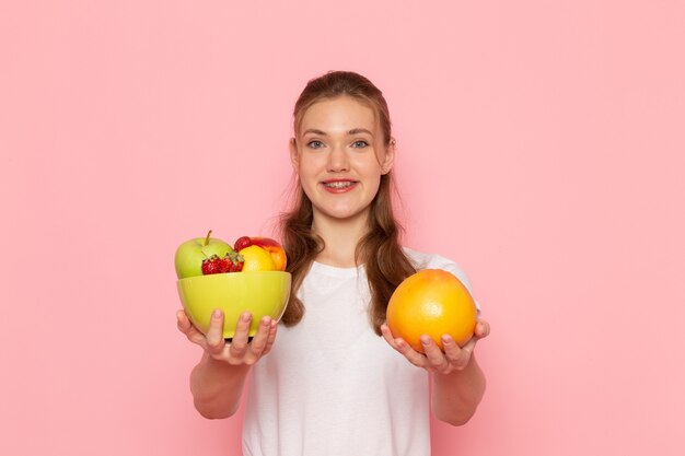 Front view of young female in white t-shirt holding plate with fresh fruits and grapefruit smiling on pink wall
