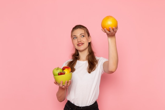 Front view of young female in white t-shirt holding plate with fresh fruits and grapefruit with smile on pink wall