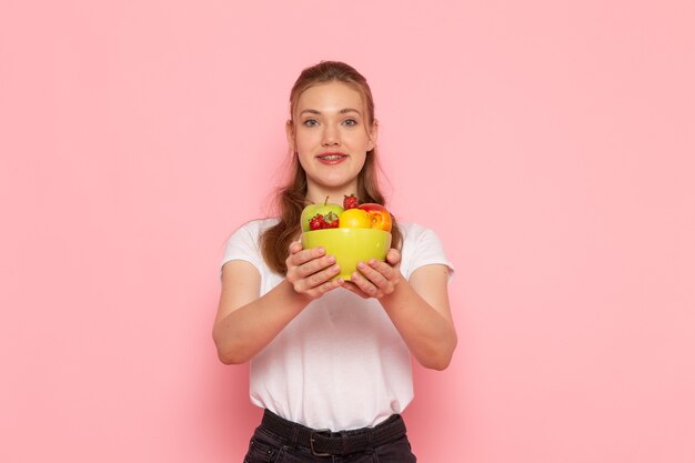 Front view of young female in white t-shirt holding plate with fresh fruits on light-pink wall