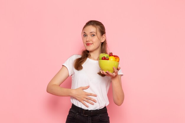 Front view of young female in white t-shirt holding plate with fresh fruits smiling on light-pink wall