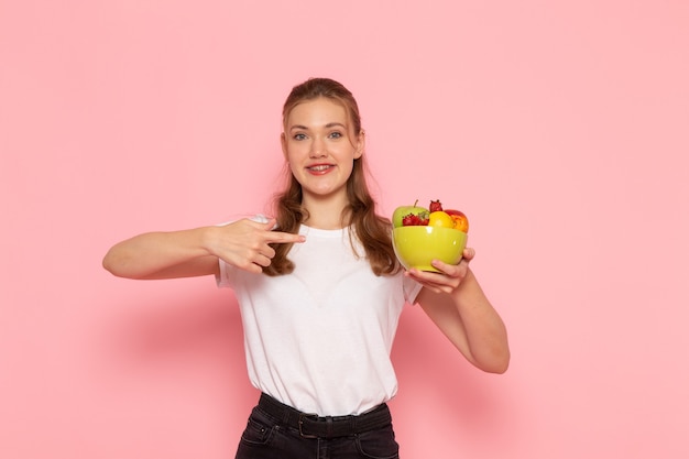 Front view of young female in white t-shirt holding plate with fresh fruits with slight smile on light-pink wall