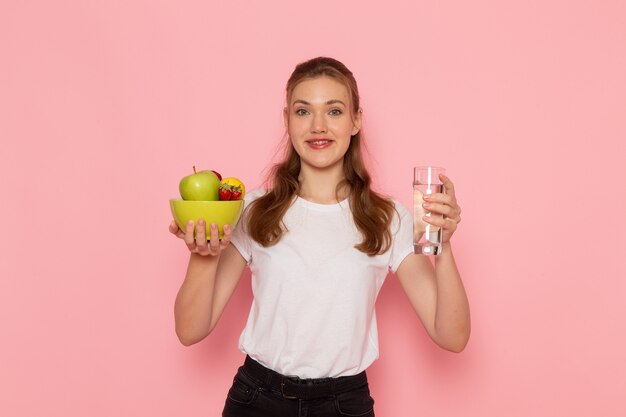 Front view of young female in white t-shirt holding plate with fruits and glass of water on pink wall