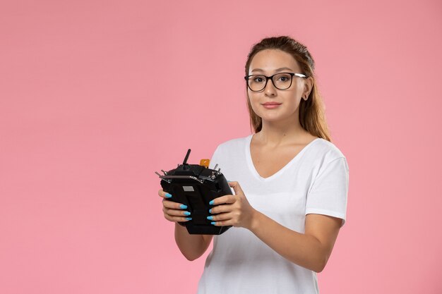 Front view young female in white t-shirt holding remote controller on the pink background 