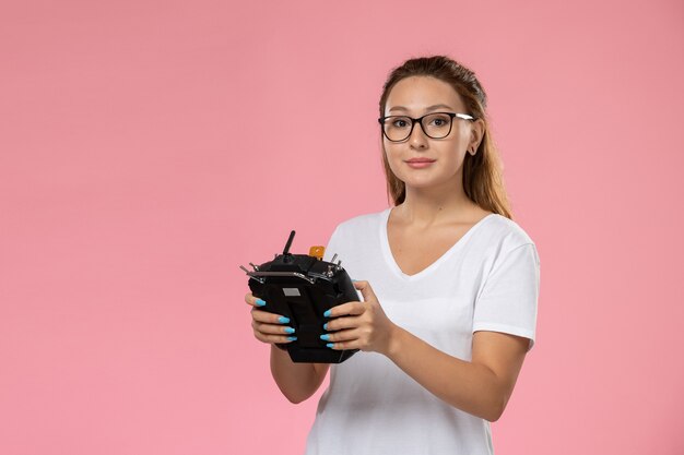 ピンクの背景にリモートコントローラーを保持している白いtシャツの正面の若い女性