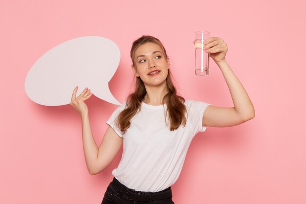 Front view of young female in white t-shirt holding white sign and glass of water smiling on the pink wall