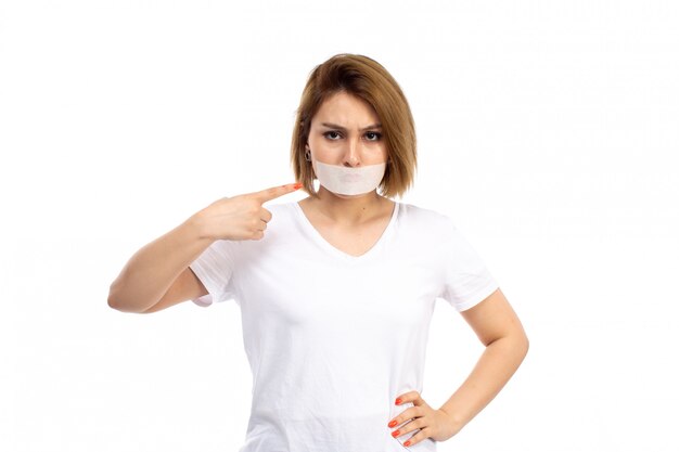 Вид спереди молодой женщины в белой футболке с белой повязкой на губах недоволен на белом