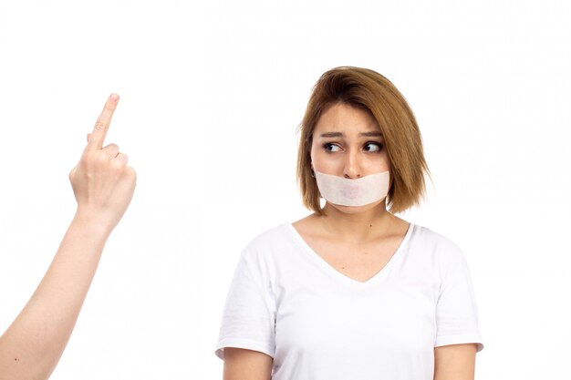 A front view young female in white t-shirt wearing white bandage around her mouth plead guilty sorry expression on the white