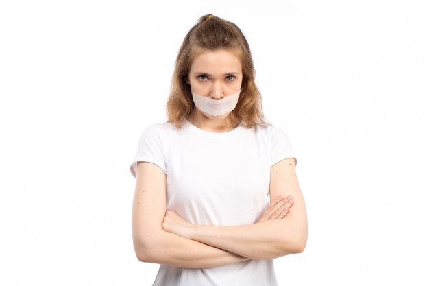 A front view young female in white t-shirt with white bandage around her mouth displeased afraid on the white